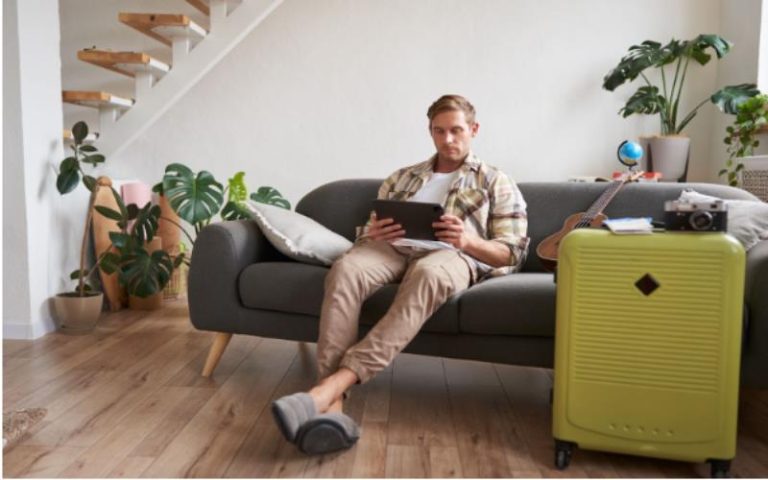 Image of young man with tablet sitting in living room with suitcase reading studying the route